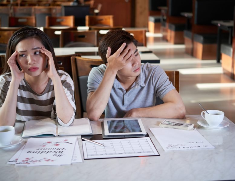 Exhausted Asian couple sitting at spacious restaurant and thinking over details of their future wedding day